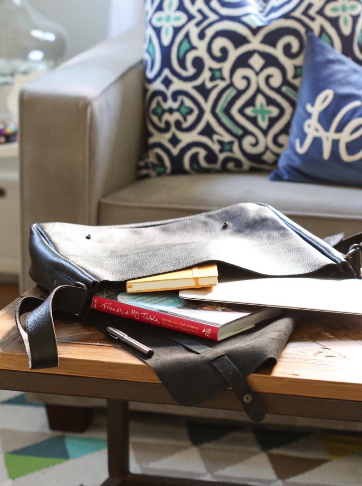 A brown messenger bag with notebooks rests on a coffee table in front of a sofa and cushions