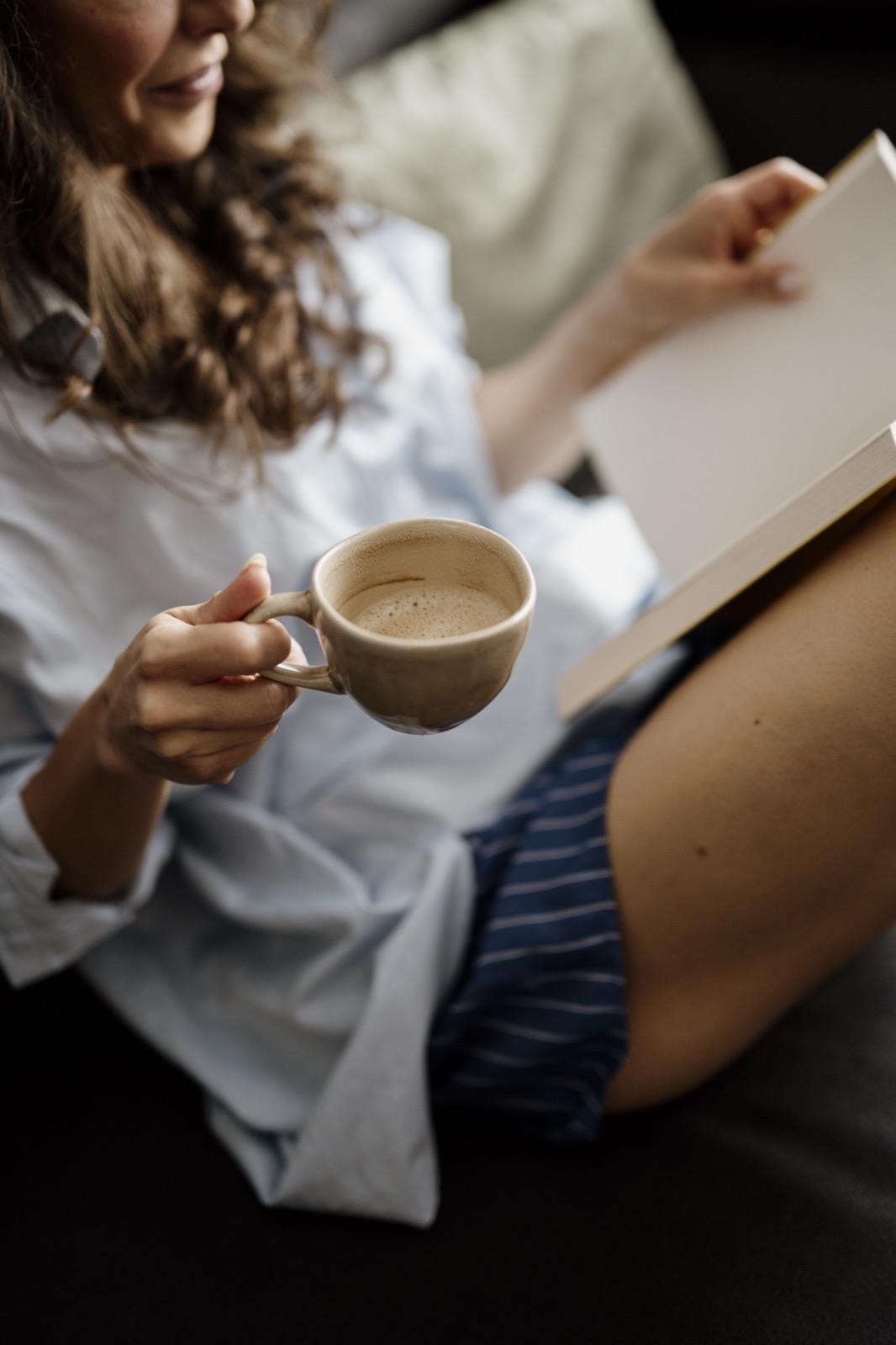 A woman seated at home holding an open book while drinking from a mug