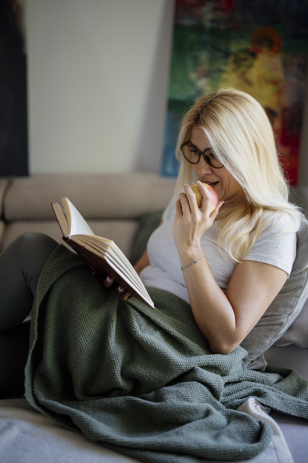 A woman reclining on a sofa with an open book, a green blanket, and an apple
